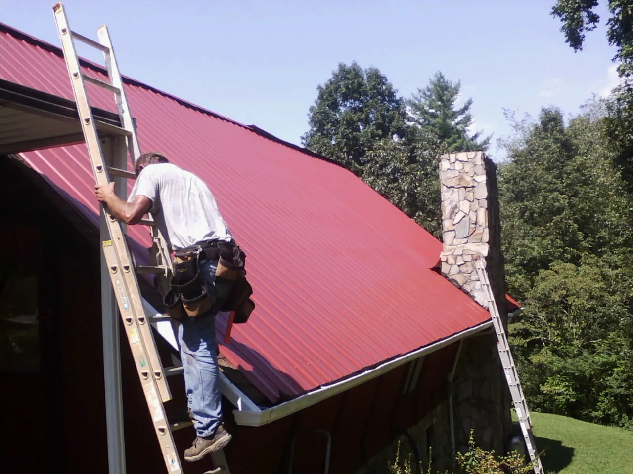 Roofer on a ladder beside a red metal roof and stone chimney — Soffit & Fascia Repair by Roofing Brothers in Provincetown